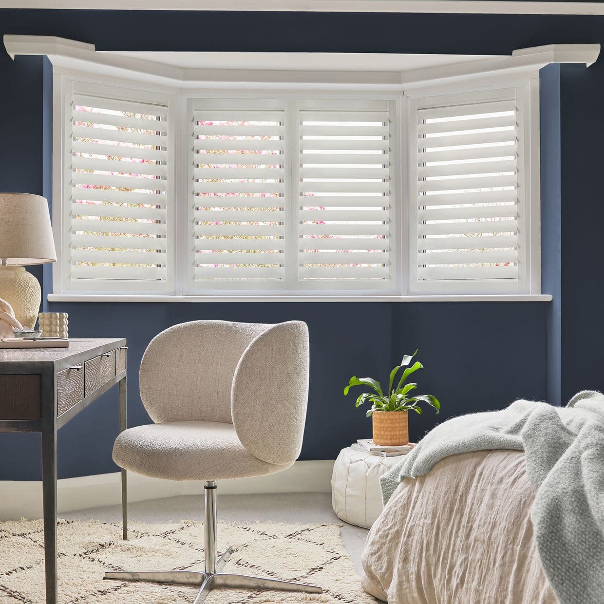 White plantation shutters covering a bay window, full-height panels with horizontal louvers â€” closed with slats partially open, filtering daylight into a navy-walled bedroom/study with beige chair, desk and bed.