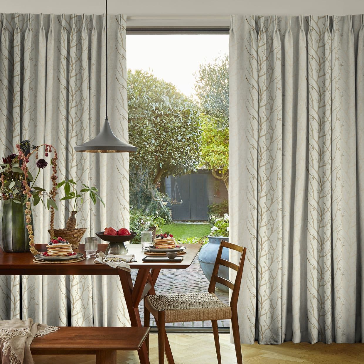 Floor-length patterned curtains, partly drawn to frame a sliding glass door, filtering daylight into a dining nook with a wooden table, pendant lamp, plates of pancakes and a green garden view.