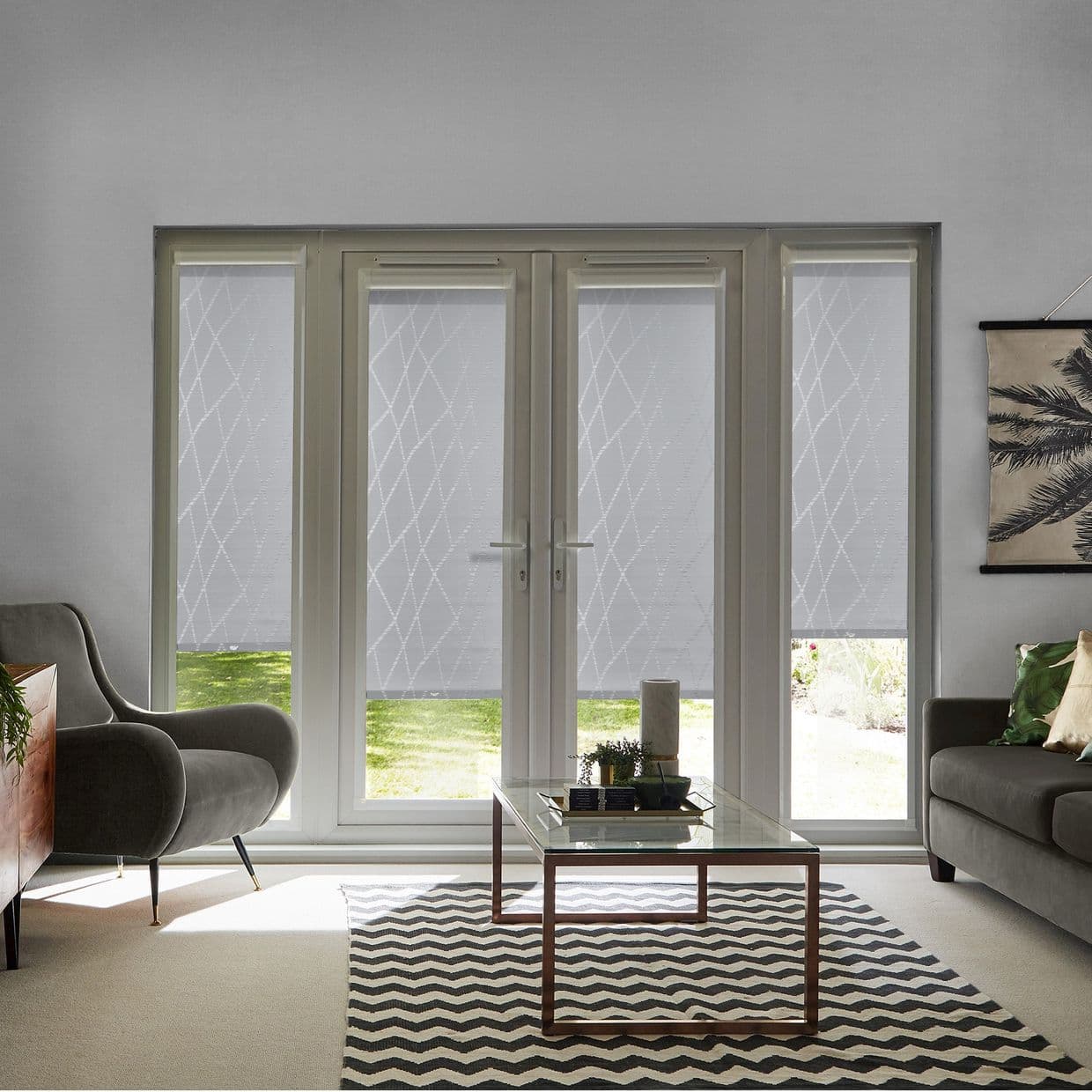 White patterned roller blinds covering four-panel French doors and sidelights, lowered and filtering daylight; modern living room with gray armchairs, glass coffee table, chevron rug, and lawn view.