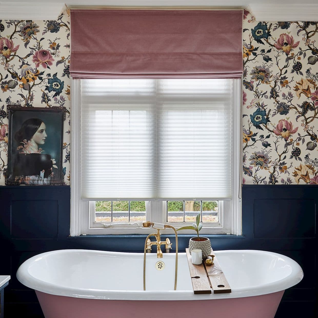 Pink Roman blind folded at top; white pleated blinds lowered, diffusing daylight over a double-sash window above a freestanding pink bathtub with dark wainscoting and floral wallpaper.