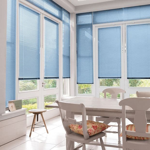 Light-blue horizontal Venetian blinds covering tall bay windows, lowered and filtering soft daylight with small bottom gaps; white dining table and chairs in a bright sunroom overlooking a green garden.