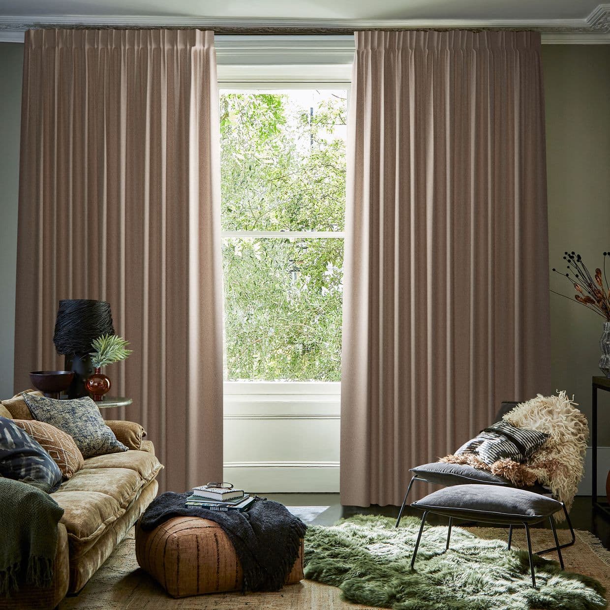Floor-to-ceiling pleated taupe drapes, split at center and framing the window, filtering soft daylight into a cozy living room with sofa, ottoman, textured rug and leafy view.