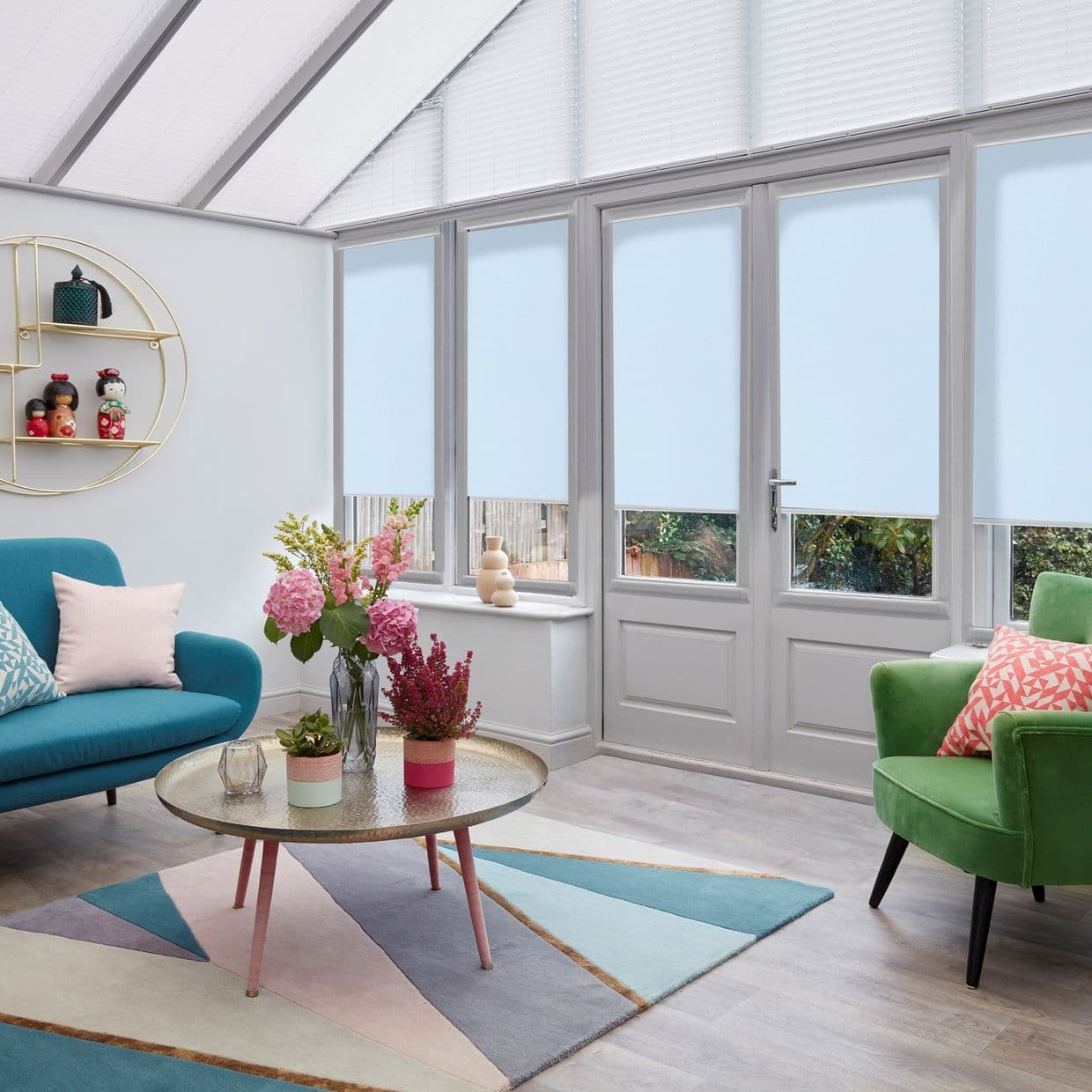 Pale blue roller blinds lowered across tall conservatory glass doors and windows, covering panes with narrow lower gaps; filtering daylight; conservatory living room with teal sofa, green armchair, floral centerpiece.