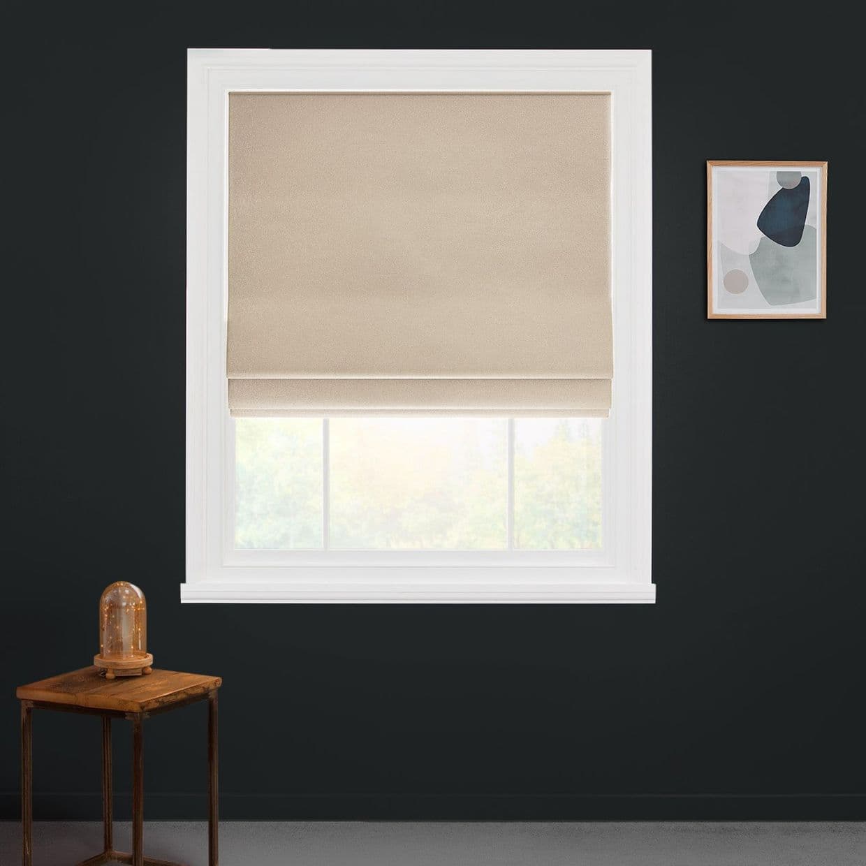 Beige Roman shade, lowered and folded at the bottom, filtering daylight; in a white-framed window on a dark charcoal wall with a small wooden table and abstract wall art.