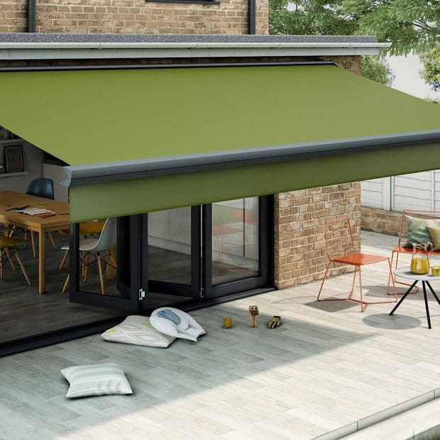An olive-green retractable fabric awning with grey casing extended over a patio, shading glass folding doors; modern brick house exterior, sunlit terrace with cushions, metal chairs and a small table.
