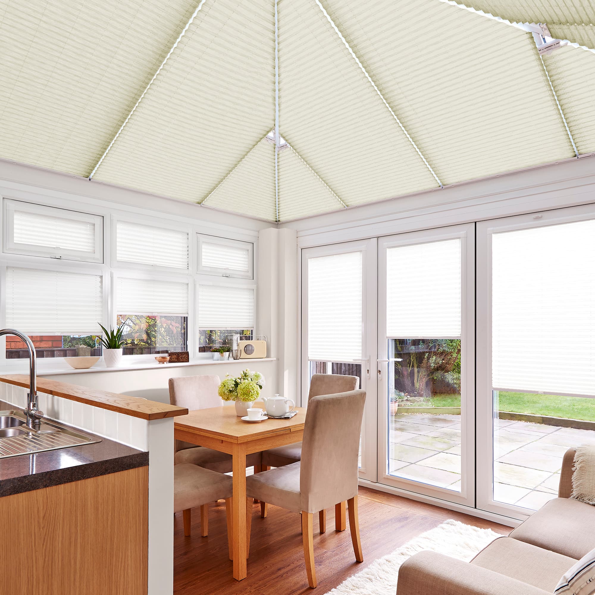 Cream pleated blinds on pitched conservatory roof and over windows and French doors, closed and diffusing bright daylight; small dining area with wooden table, beige chairs, sink and garden view.