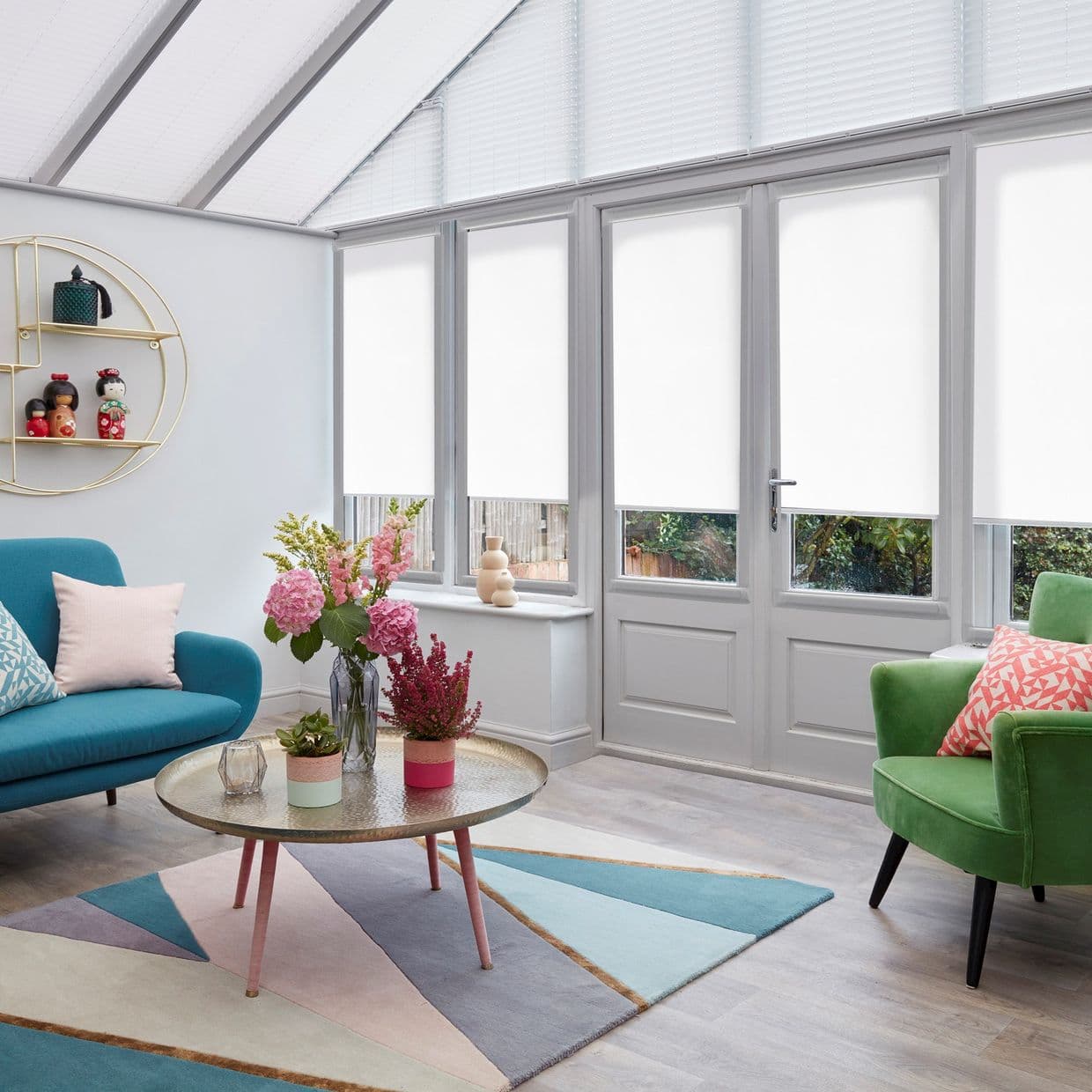 White roller blinds lowered over tall conservatory windows and door, filtering daylight and obscuring upper views; bright sunroom with teal sofa, green armchair, floral centerpiece and geometric rug.