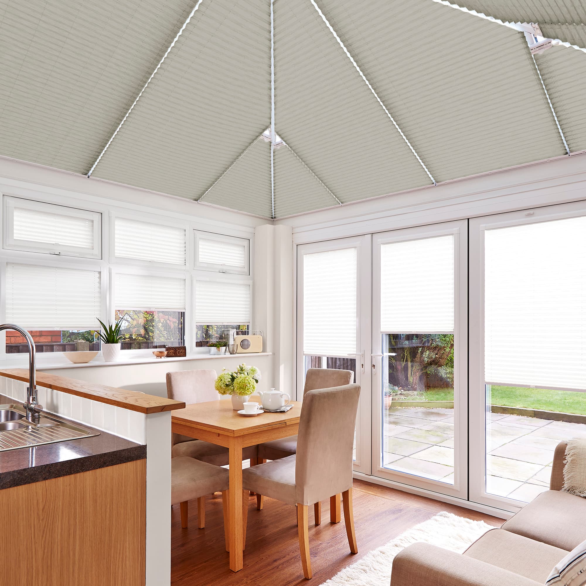 Pleated roof blinds across pitched conservatory ceiling, pale beige with visible ribs, fully lowered and diffusing daylight; kitchen-dining conservatory with wooden table, upholstered chairs, sink and glazed doors to patio.