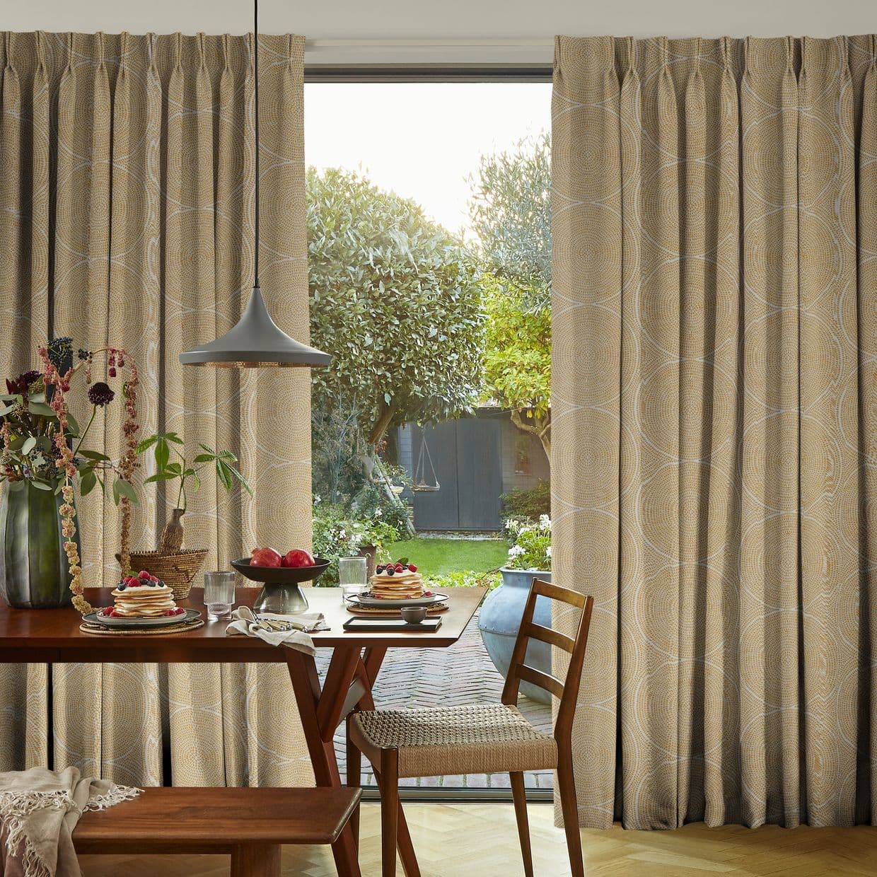 Floor-length beige pleated curtains with circular pattern, parted centrally to reveal sliding glass door. They frame daylight over a dining table set for breakfast and a garden beyond.