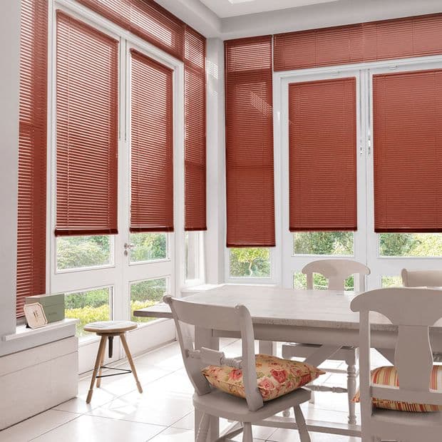 Red horizontal Venetian blinds across floor-to-ceiling bay windows and glass door, fully lowered and filtering daylight; bright white dining nook with wooden table, chairs and floral cushions, garden visible.