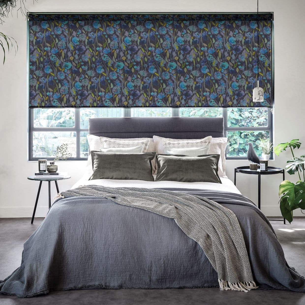 Floral-patterned roller blind (blue flowers on dark background), lowered across the upper window, filtering daylight; bedroom with a grey-upholstered bed, layered bedding, bedside tables, plants and a pendant light.