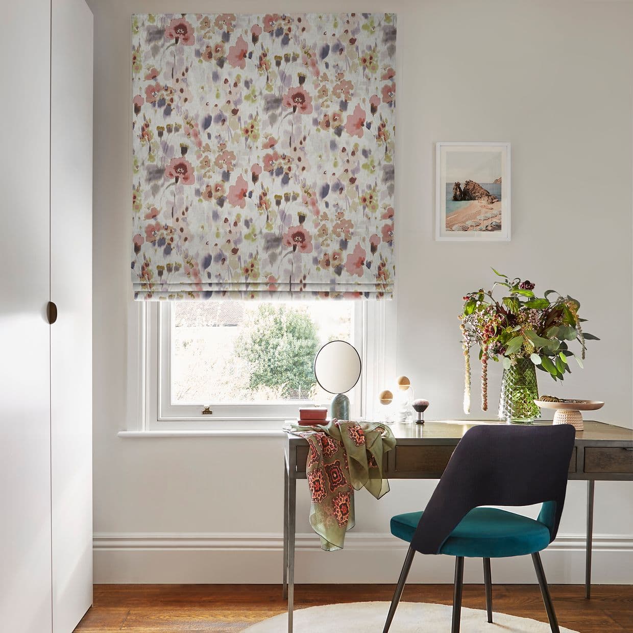 Floral Roman blind with pastel watercolor poppy pattern, partially lowered and filtering soft daylight over a wooden desk setup â€” mirror, vase of flowers, teal chair in a neutral-toned home office.