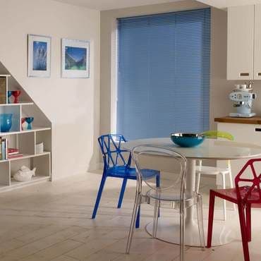 Blue horizontal blinds, fully lowered and closed, fine slats filtering daylight over a recessed window. In a bright modern dining-kitchen with a round glass table, colorful transparent chairs and shelving.