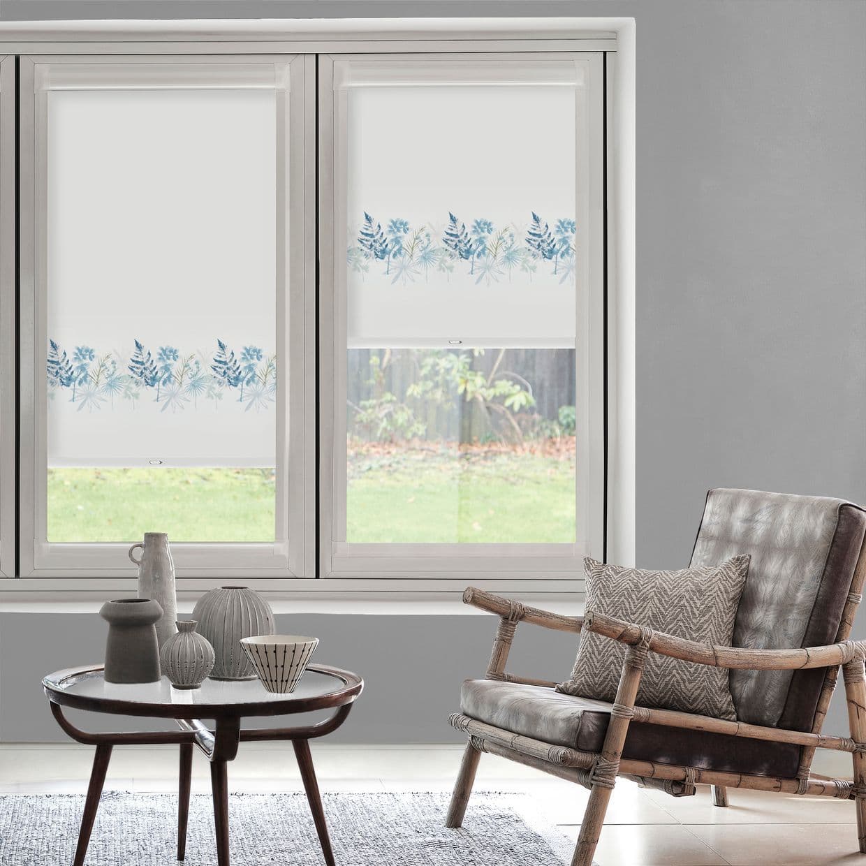 White roller blinds with blue botanical border, lowered halfway across twin windows, filtering soft daylight; living room seating with rattan armchair, side table with ceramics, neutral gray walls, garden view.