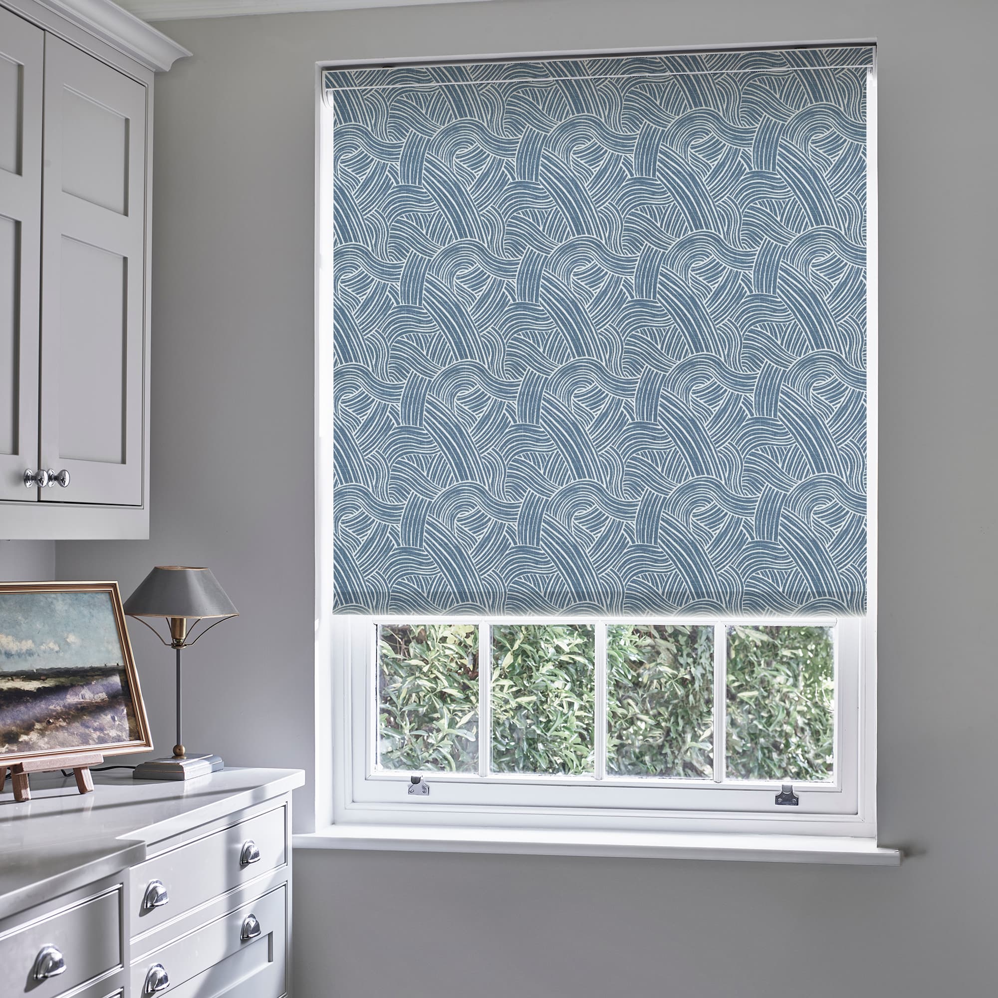 Blue roller blind with white braided-line pattern is fully lowered, filtering light; in a white-framed sash window of a grey-walled room with white chest, lamp and framed painting, greenery outside.
