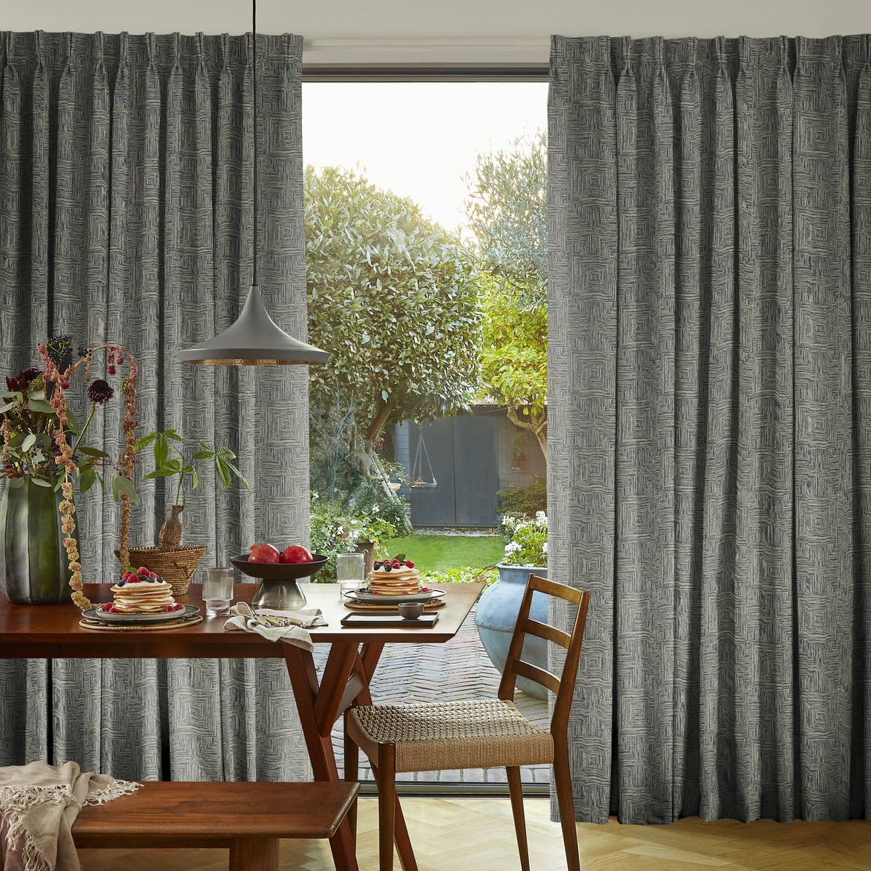Floor-to-ceiling pleated grey patterned curtains, hung in soft folds and parted centrally; drawn partly open, admitting daylight and a garden view; wooden dining table set for breakfast beneath a pendant lamp.