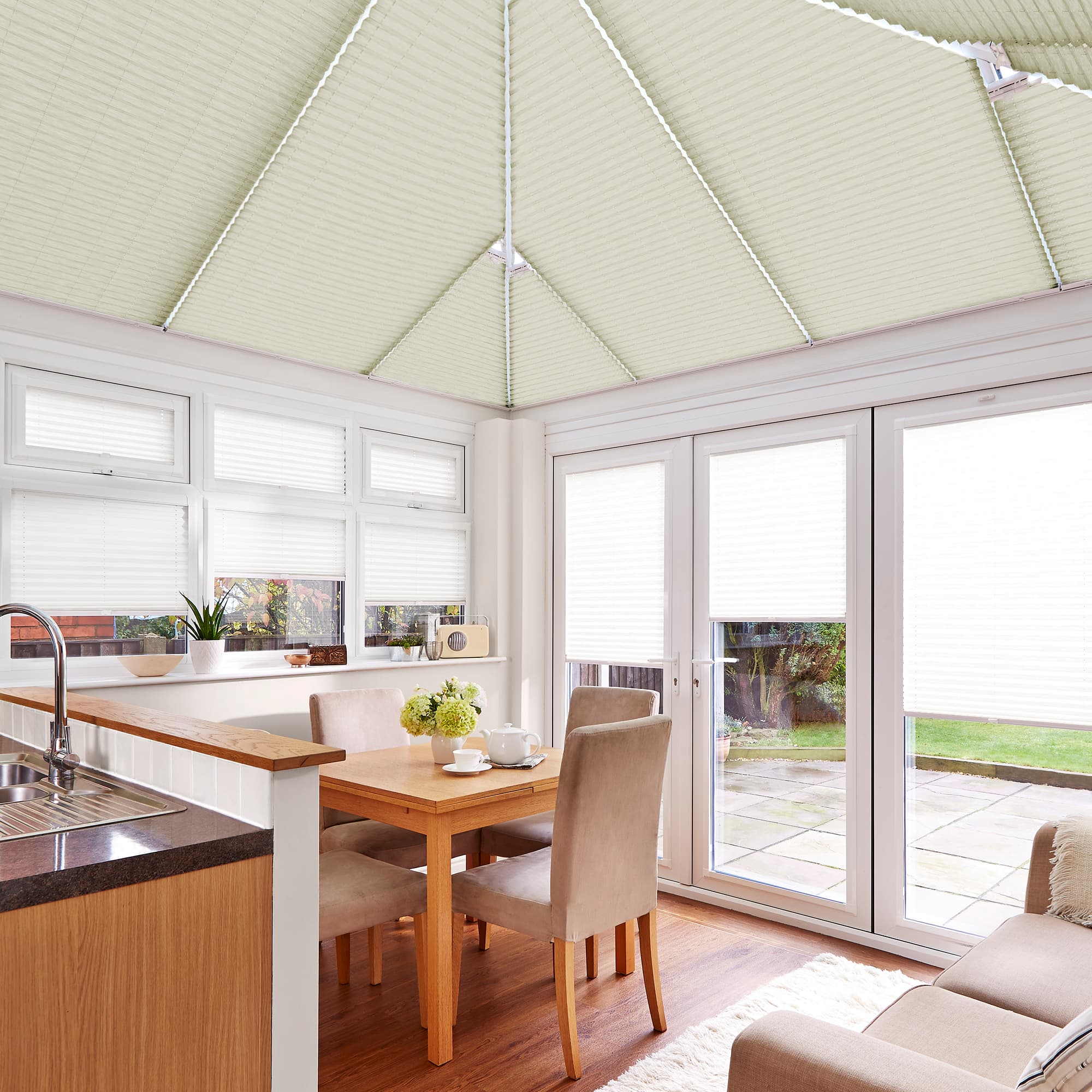 Pleated white cellular blinds on the pitched conservatory roof and covering windows and glass doors, closed and diffusing daylight; dining-kitchen conservatory with wooden table, beige chairs, and garden view.