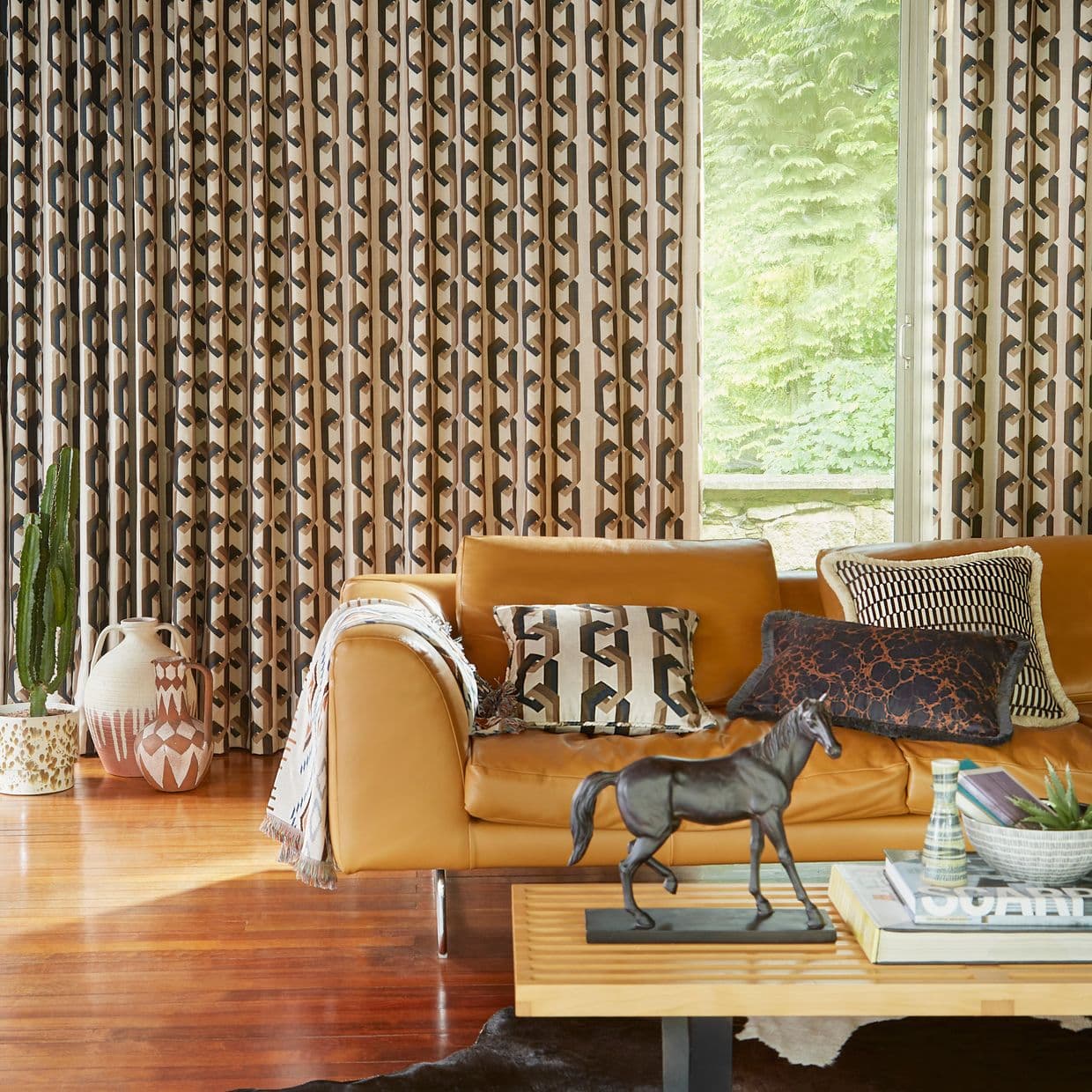 Full-height pleated geometric curtains (tan, brown, black), mostly drawn with a narrow opening; filtering daylight into a living room with tan leather sofa, wooden floor, coffee table and garden view.