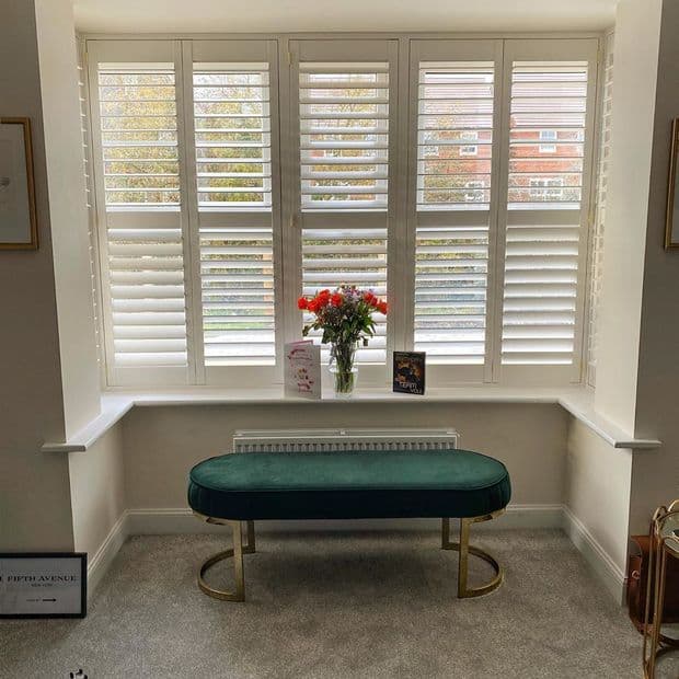 White plantation shutters cover a bay window, louvers partially tilted, filtering daylight; bright sitting alcove with a green velvet bench on gold legs and a vase of red flowers.