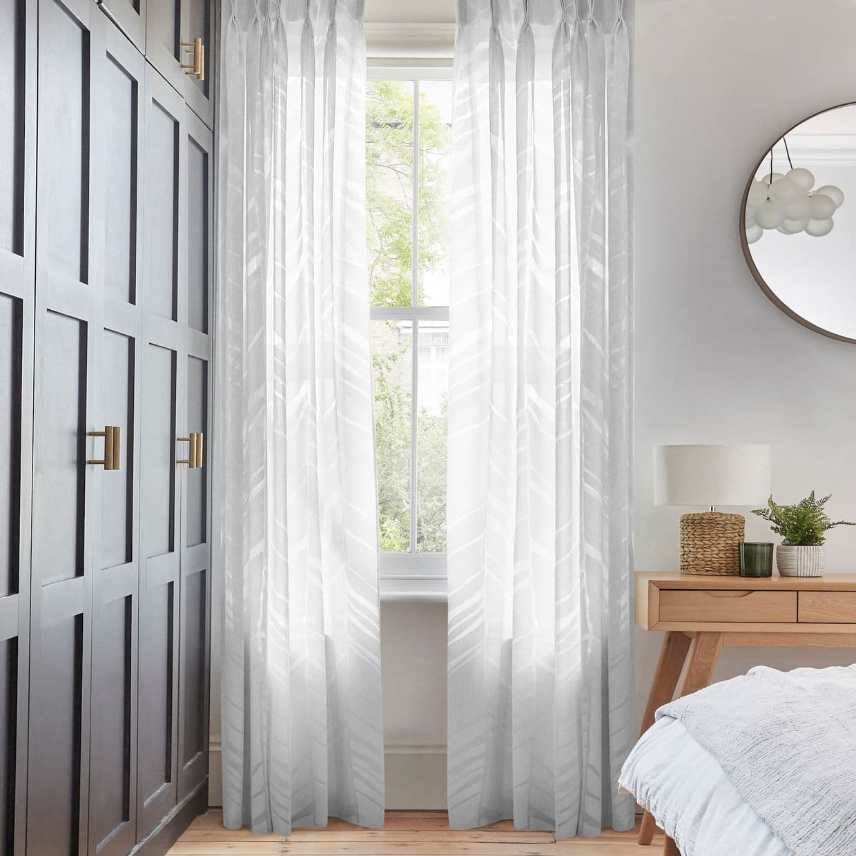 Sheer patterned curtains framing a tall sash window, filtering soft daylight into a bedroom corner with dark paneled wardrobe, wooden bedside table, lamp, mirror, and green outside view.