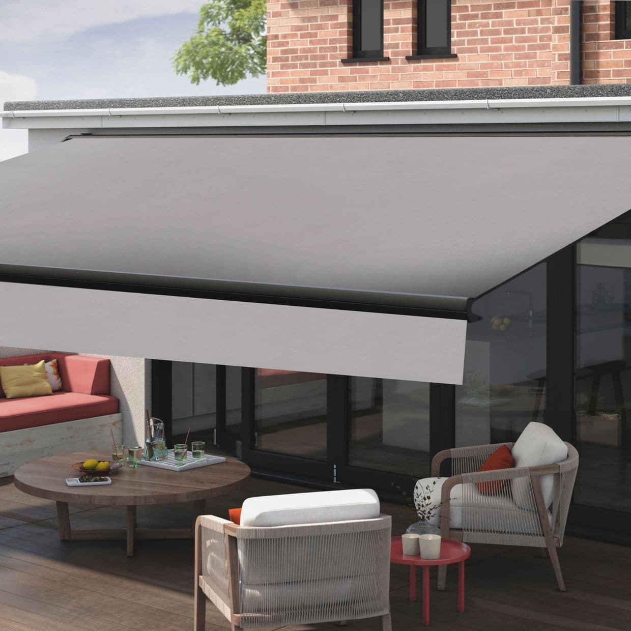 Retractable gray fabric awning (fascia-mounted, black front bar), fully extended and shading the area; outdoor wooden-deck patio with wicker chairs, low round table, glass doors and brick upper wall, daytime.
