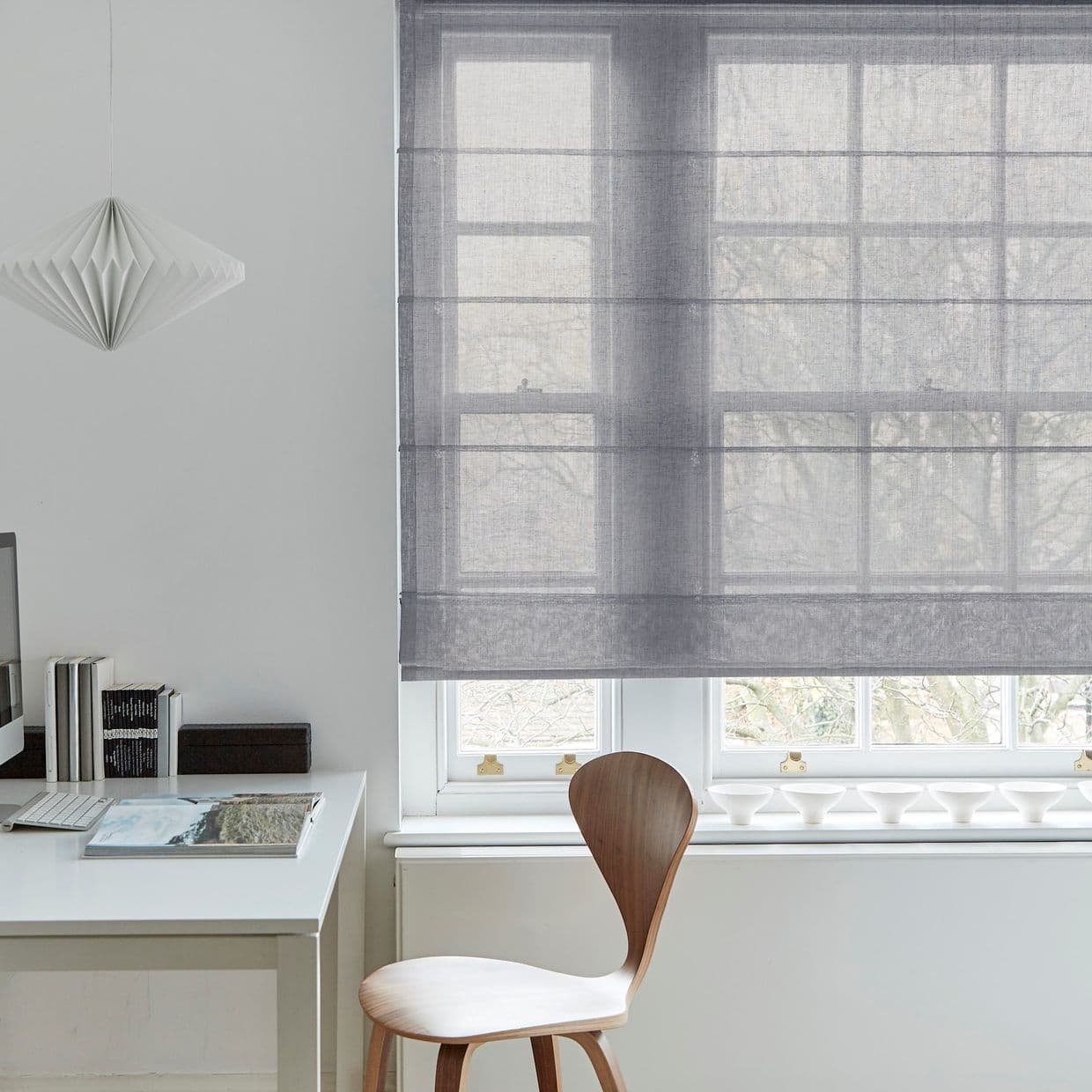 Sheer grey Roman blind, lowered across a multi-pane window with horizontal folds, filtering soft daylight; minimalist home office with white desk, wooden chair, pendant lamp, and bare tree branches outside.