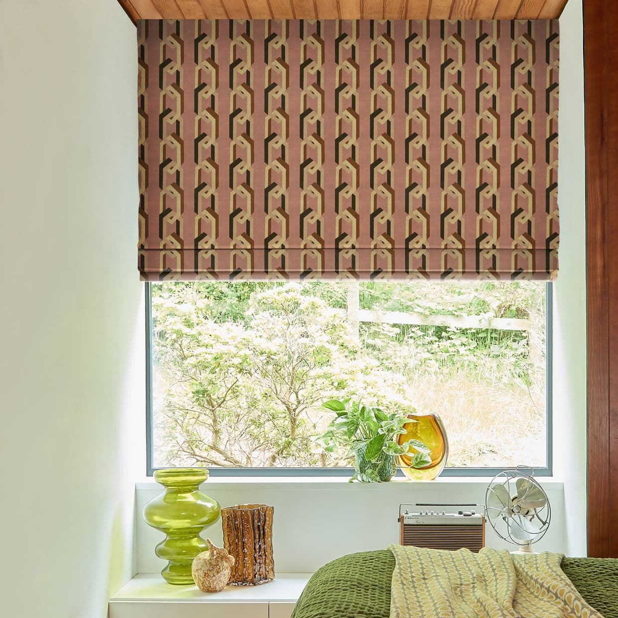 Geometric Roman blind (mauve, beige) lowered over the upper window, partly blocking light; bedroom sill with plants, green and amber glass vases, a small fan, and a leafy garden view.
