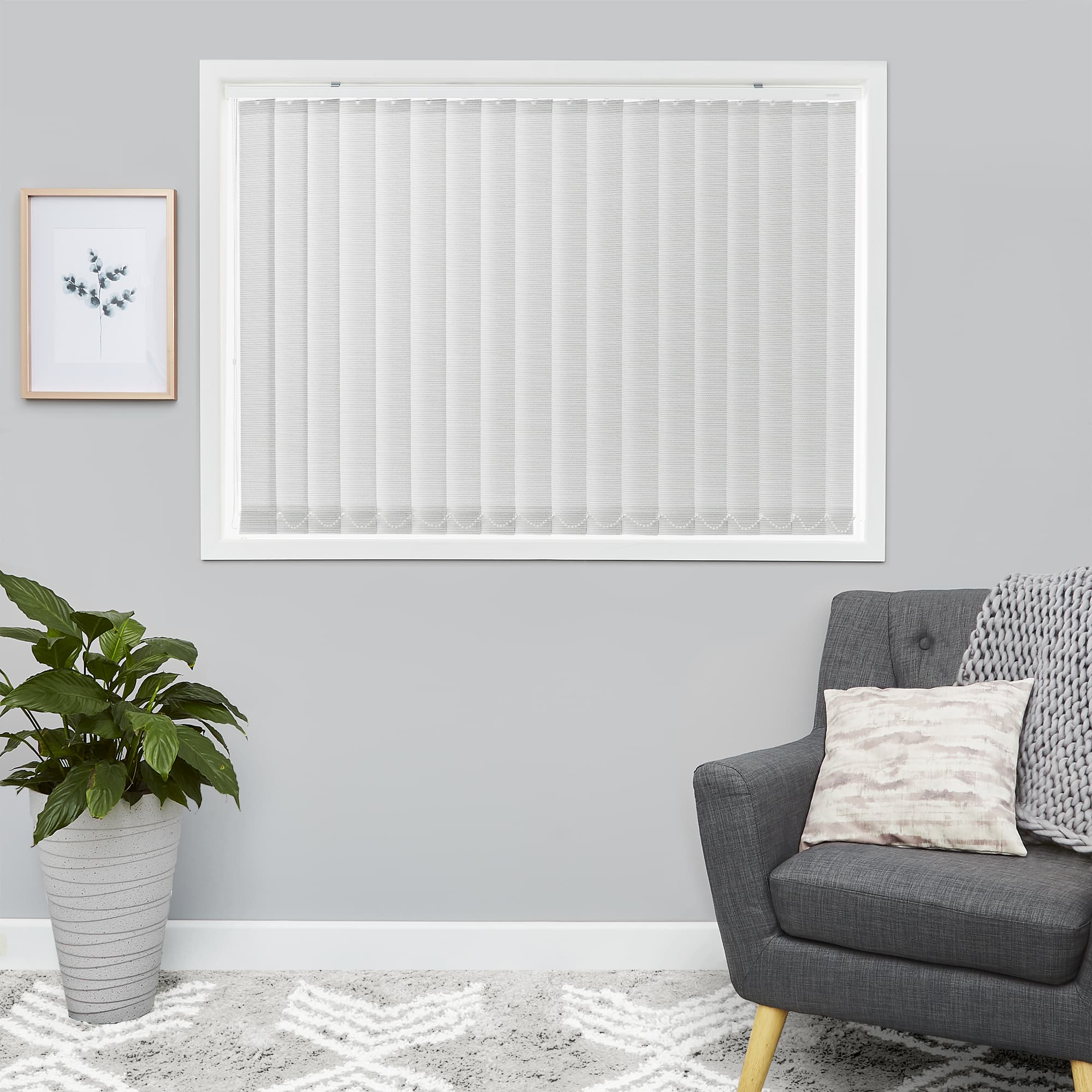 White vertical blinds fill a rectangular window â€” evenly spaced slats. They are fully closed, softly diffusing daylight. Neutral grey living room with charcoal armchair, potted plant, and framed print.