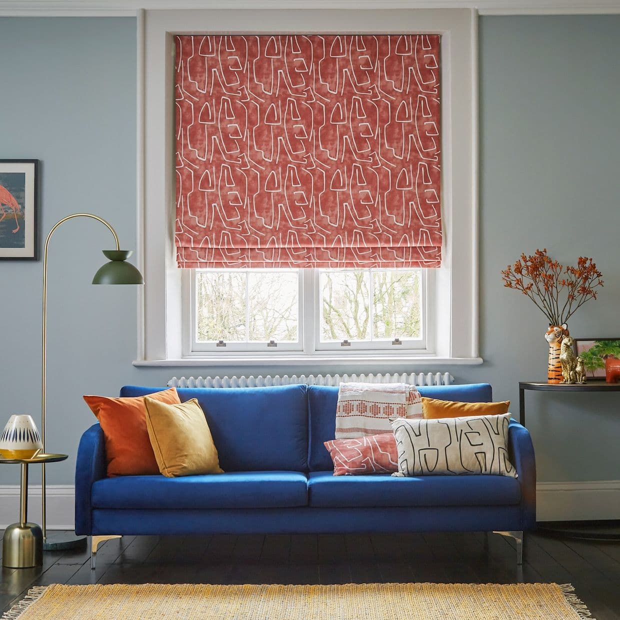 A terracotta patterned Roman blind pulled down over a double window, filtering daylight above a cobalt sofa with colorful cushions, floor lamp and side table in a lit living room.