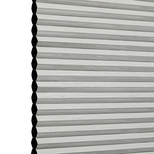 Honeycomb (cellular) blind panel at left edge, white-gray pleated horizontal cells with black hexagonal cell ends; lowered and closed, filtering diffuse light; studio-lit close-up against plain white background.