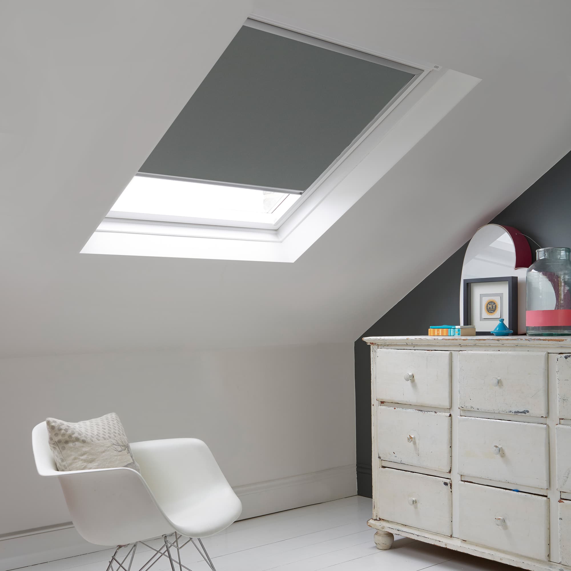 Room with white eaves wall, dark blue-grey wall to right with a shabby chic white drawer unit, a contemporary white chair, white painted floorboards and a skylight blind in steel.