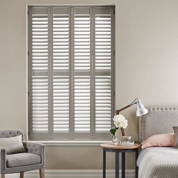 White four-panel louvered plantation shutters covering a tall rectangular window, slats tilted to filter daylight; bedroom setting with upholstered bed, side table with lamp, flowers and a tufted armchair.