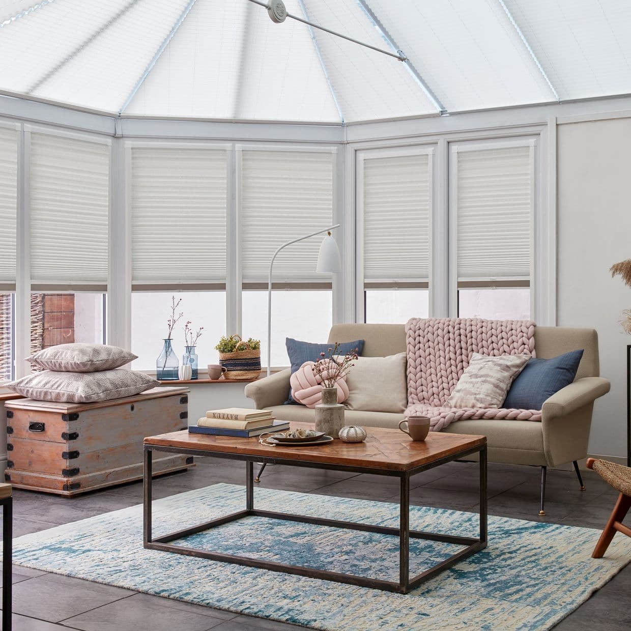 White honeycomb pleated blinds covering bay windows and conservatory roof, filtering soft daylight; cozy sunroom with a sofa, chunky knit throw, wooden coffee table and blue patterned rug.