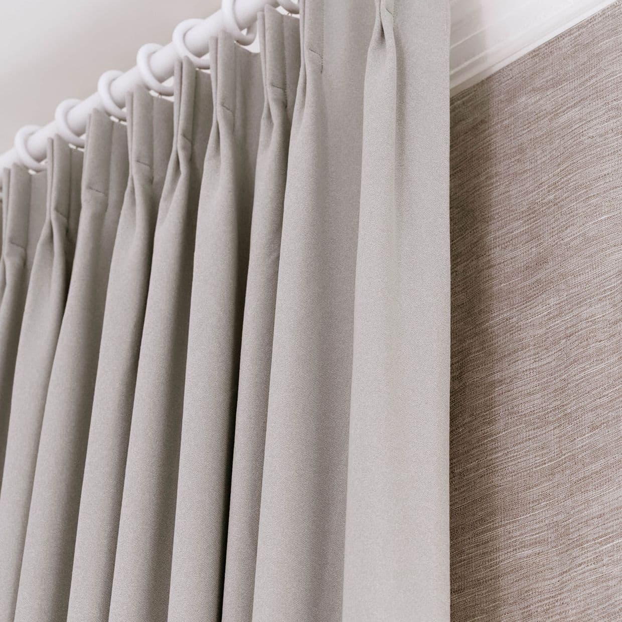 Pleated grey fabric curtains hung from a white ring-top rod, tightly closed and evenly pleated; they obscure a textured beige wall covering beside them, bathed in soft, diffused indoor light.