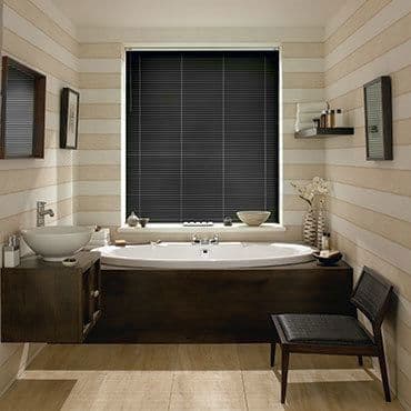 Black horizontal blinds, fully closed with thin slats covering a central window; blocking daylight; above a white tub in a warm wood-paneled bathroom with sink, shelf and chair.