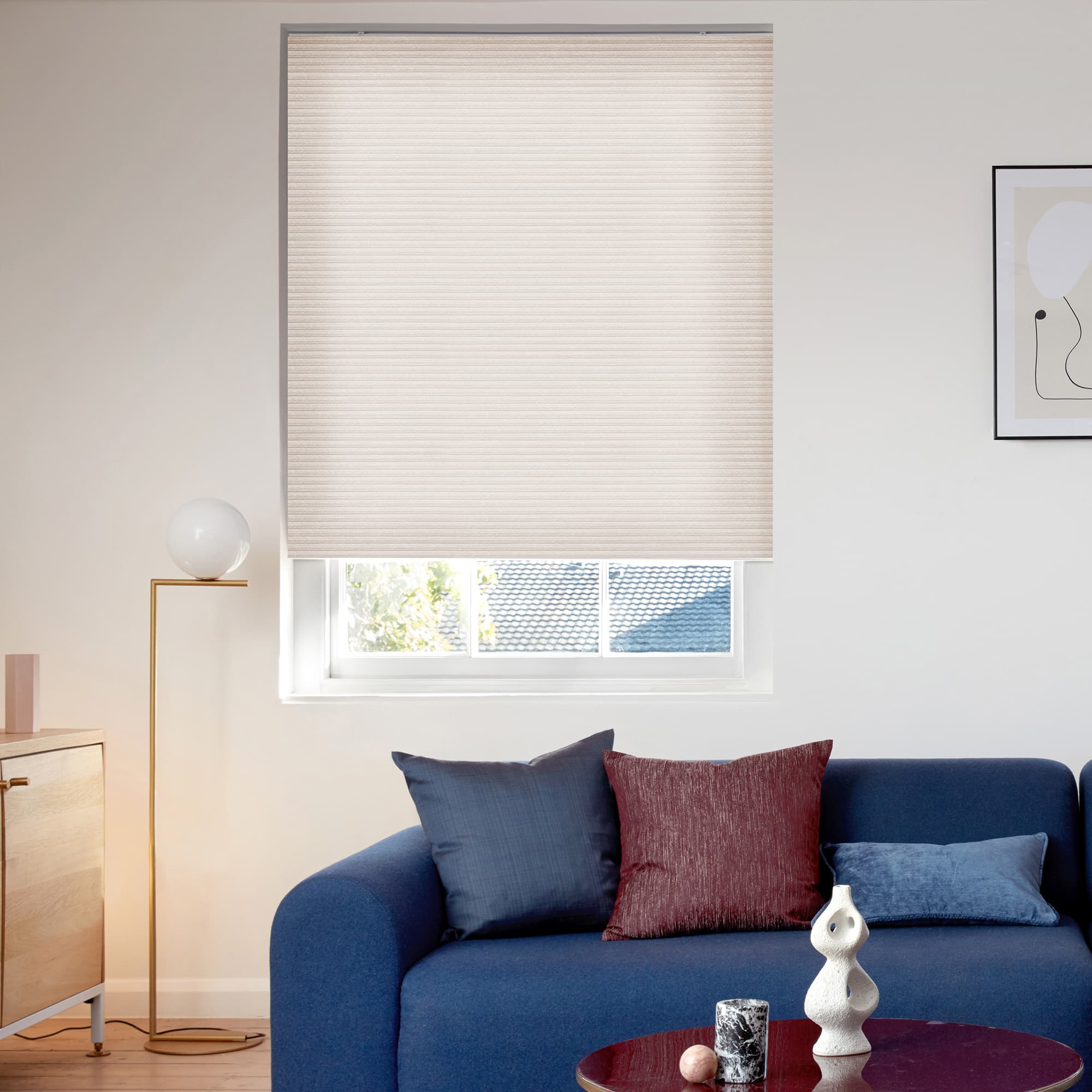 A beige cellular (honeycomb) roller blind lowered over a window, filtering soft daylight; living room with blue sofa and cushions, floor lamp and sideboard, glimpse of roof and tree outside.