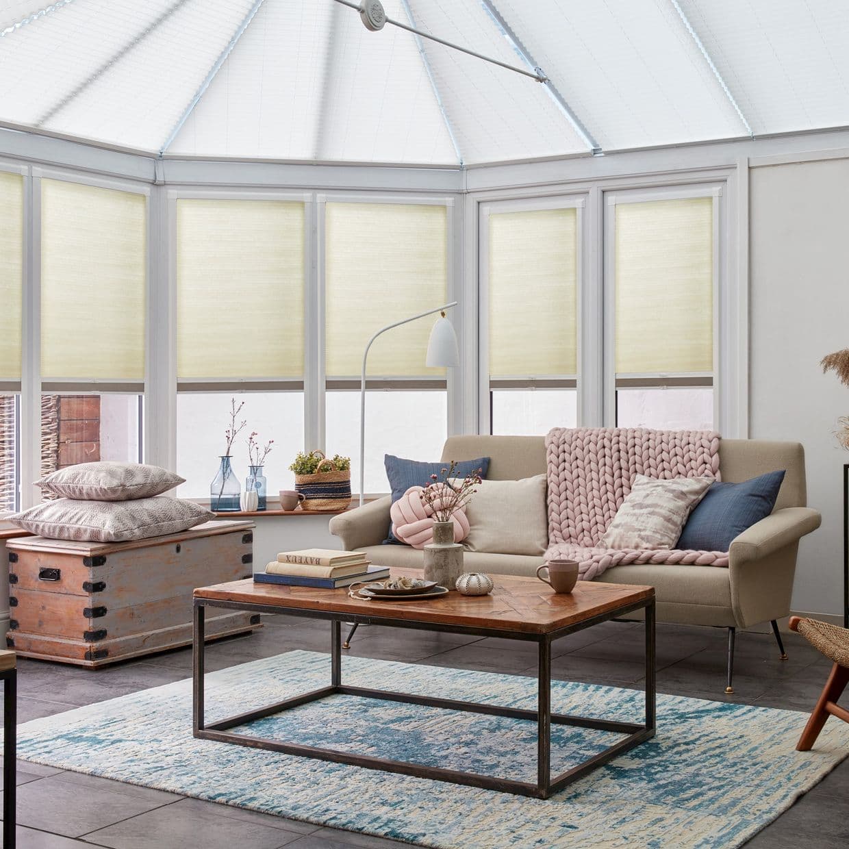 Cream pleated cellular blinds, lowered across tall conservatory windows, filtering soft daylight; set in a bright sunroom with vaulted glass roof, beige sofa, wooden coffee table and blue rug.