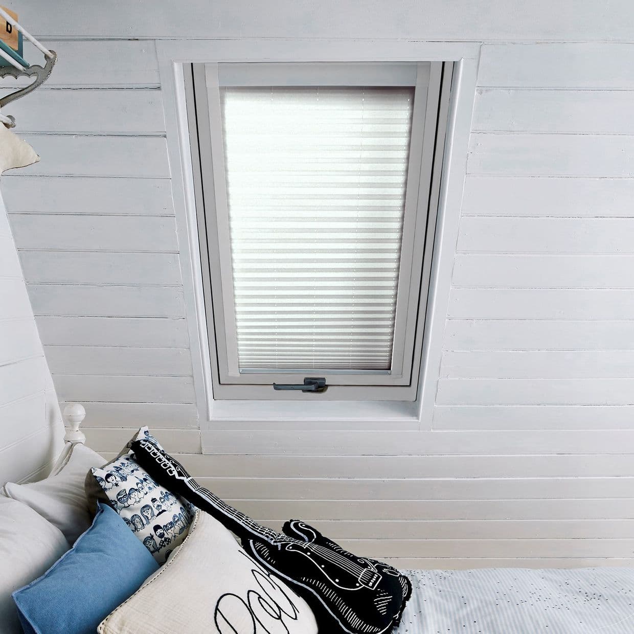 White pleated blind fully closed over a rectangular skylight, softly diffusing daylight; set in a bright white shiplap attic bedroom above a bed corner with blue, patterned and guitar-shaped cushions.
