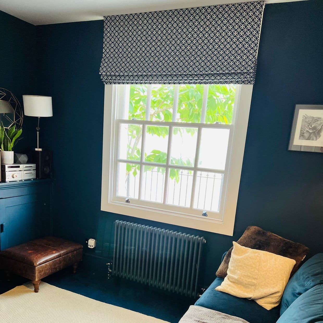 Roman fabric shade with navy-and-white geometric pattern, mounted above the sash window and folded at the top; lowered partially, filtering bright daylight; cozy dark-teal living room with radiator, sofa, lamp.