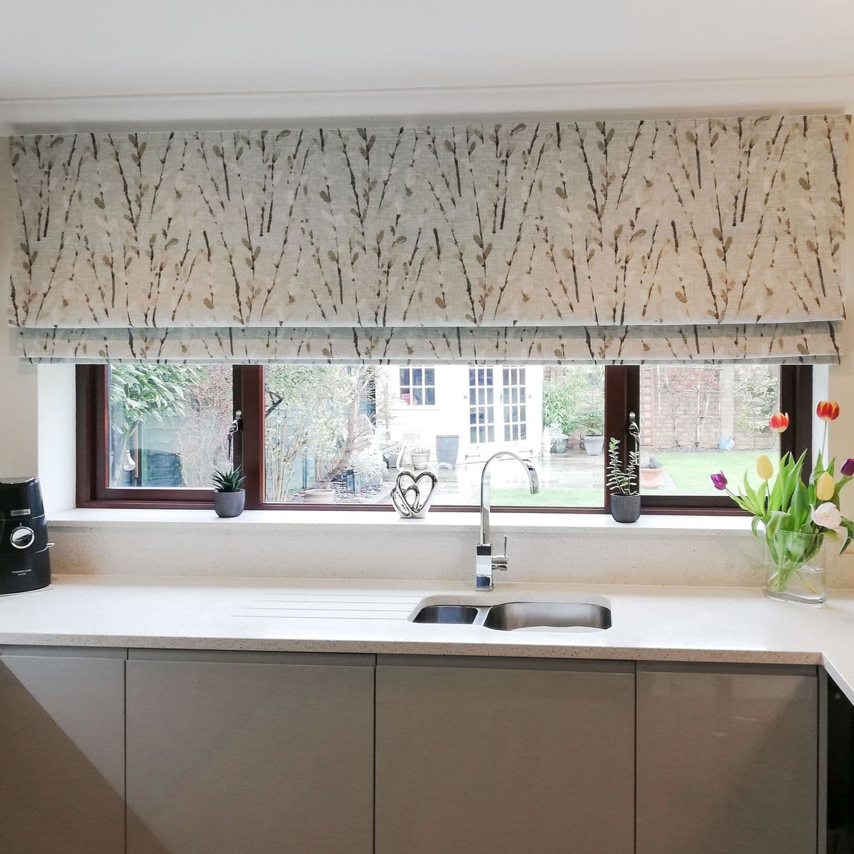 Fabric Roman blind with subtle twig pattern, lowered and partly folded, filtering daylight over a kitchen sink and countertop; stainless faucet, small potted plants and tulips, view of garden.