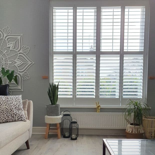 White plantation shutters covering a tall four-panel window, horizontal louvers partially open and filtering bright daylight; in a light grey living room with sofa, potted plants, wall art and lanterns.