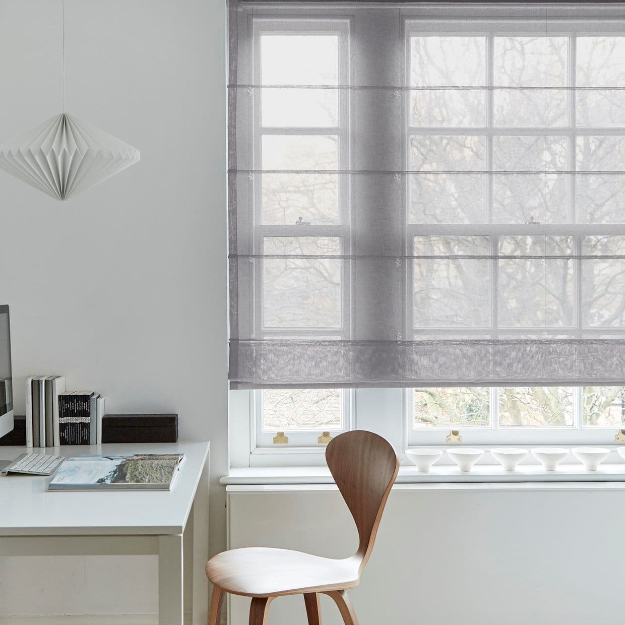 Sheer grey Roman blind, lowered with horizontal folds, filtering pale daylight over a multiâ€‘pane window in minimalist home office with white desk, wooden chair, pendant lamp and bare trees outside.