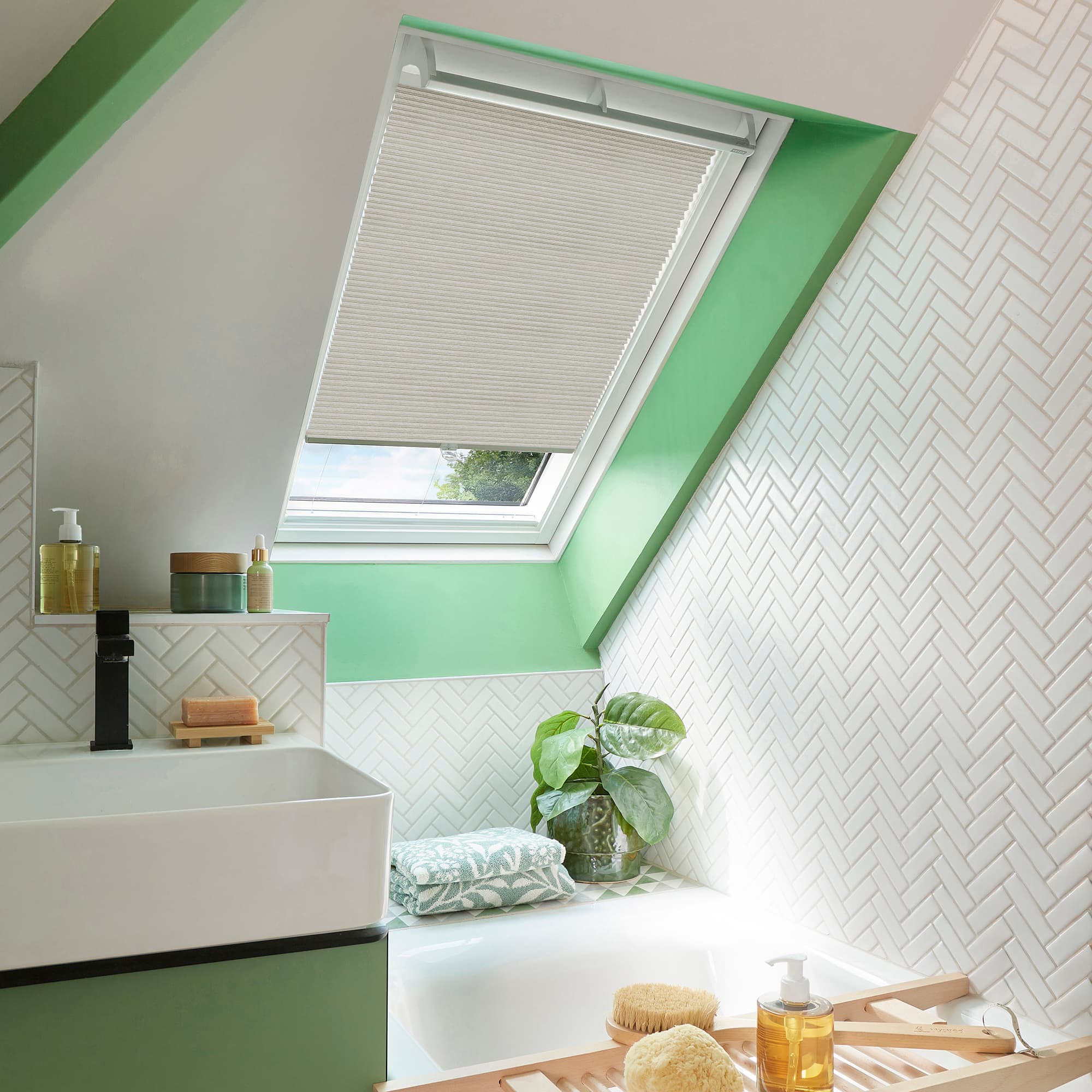Pleated beige skylight blind, partially lowered across a slanted roof window, filtering daylight; set above a green-accented attic bathroom with white herringbone tiles, sink, bathtub, and a potted plant.