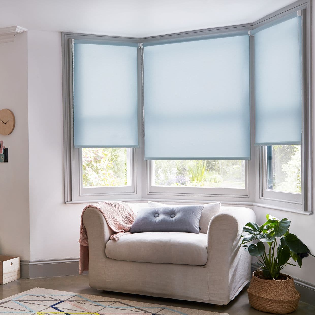 Light blue roller blinds on a three-panel bay window, lowered and semi-opaque, filtering soft daylight into a cozy living room with a beige armchair, cushion, throw and potted plant.