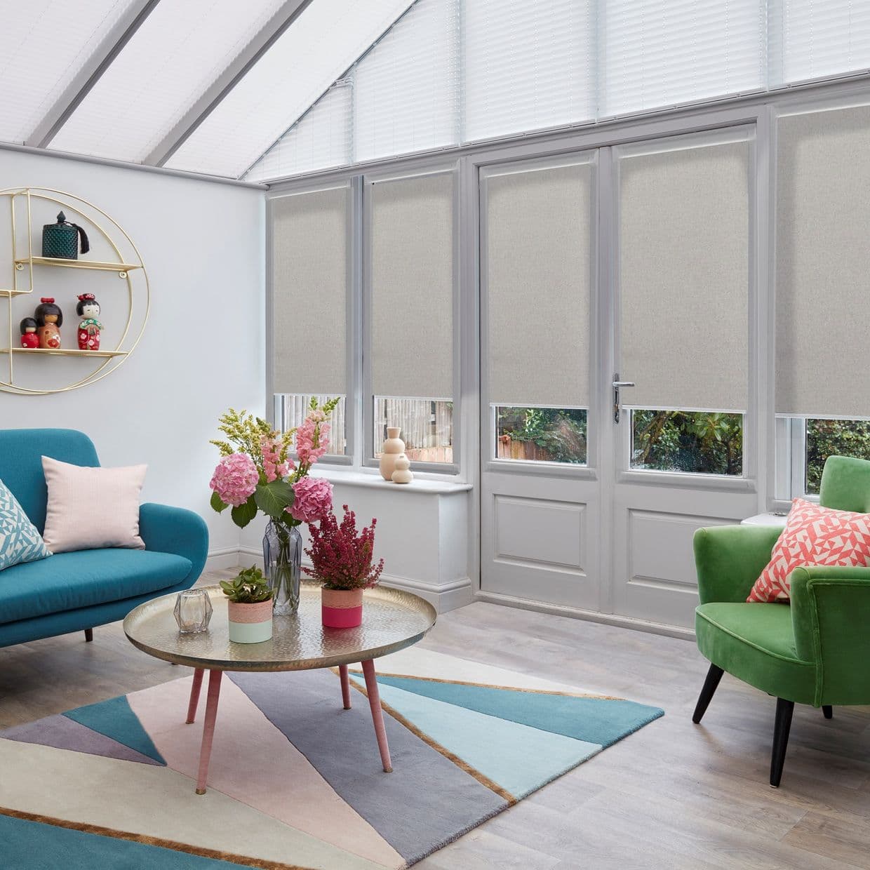 Beige roller blinds across tall glazed doors, lowered to cover most glass but leaving narrow bottom panes visible, filtering daylight and providing privacy in a bright sunroom with colorful chairs.