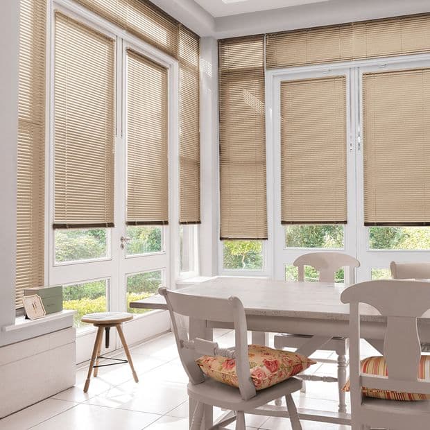 Beige horizontal Venetian blinds covering tall French windows, lowered and filtering daylight; kitchen-dining nook with white table and chairs, floral cushions, tiled floor and green garden visible through lower glass.