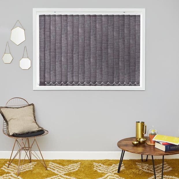 Vertical blinds, closed over a rectangular window, textured gray fabric with scalloped white chain at bottom, light-blocking; on a gray living room wall above a chair, rug and coffee table.