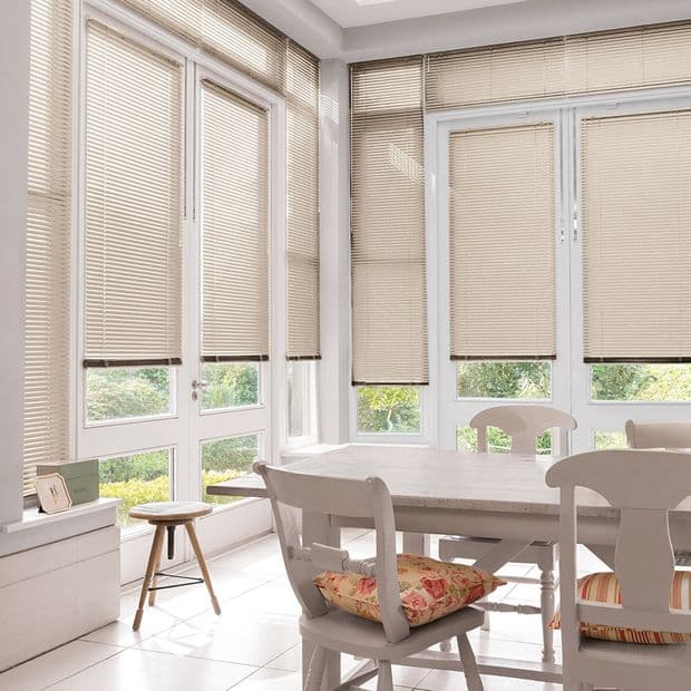 Beige horizontal blinds covering tall bay windows, lowered with slats slightly tilted, filtering soft daylight into a bright dining nook with white table, chairs, floral cushions, and a garden view.