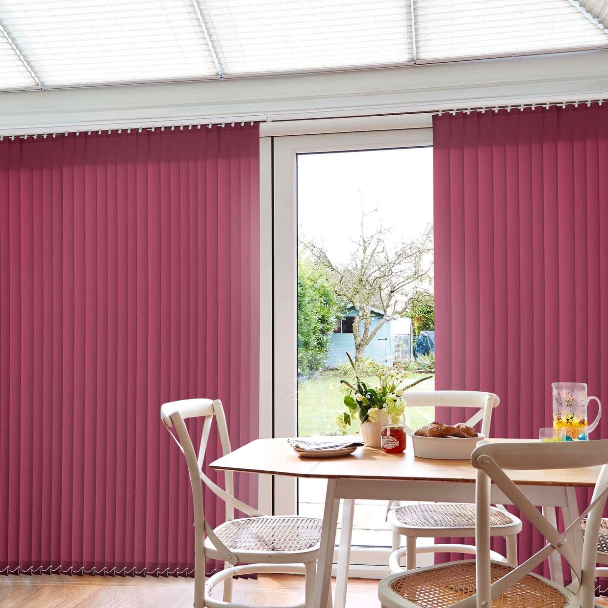 Vertical magenta fabric blinds flank a sliding glass door, partially drawn to leave the center panel clear and filter daylight; small dining table and white chairs in foreground, backyard visible.