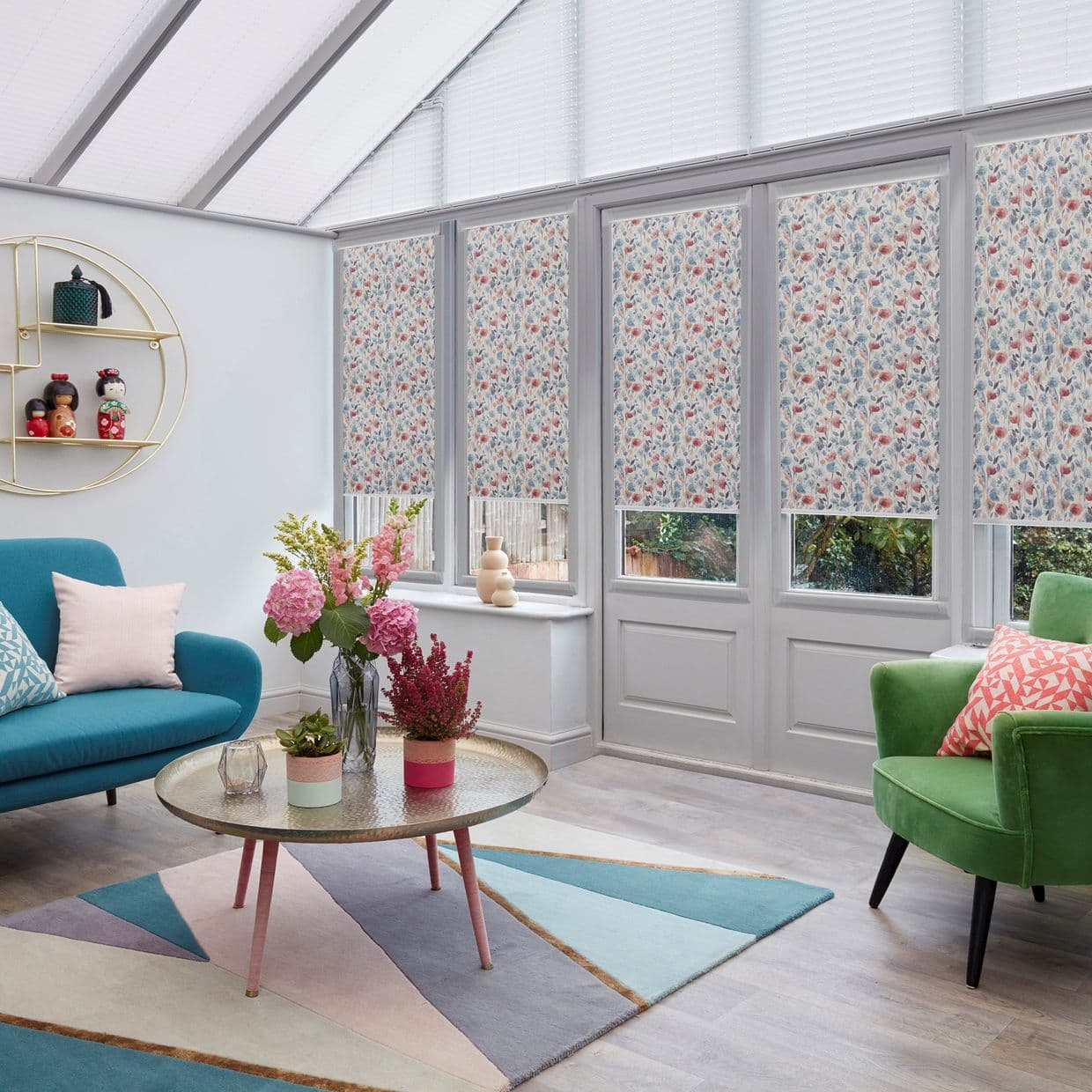 Floral roller blinds on tall conservatory windows and glazed doors; lowered, partially blocking and filtering daylight; bright sunroom with skylight, teal sofa, green armchair, coffee table and flowers.